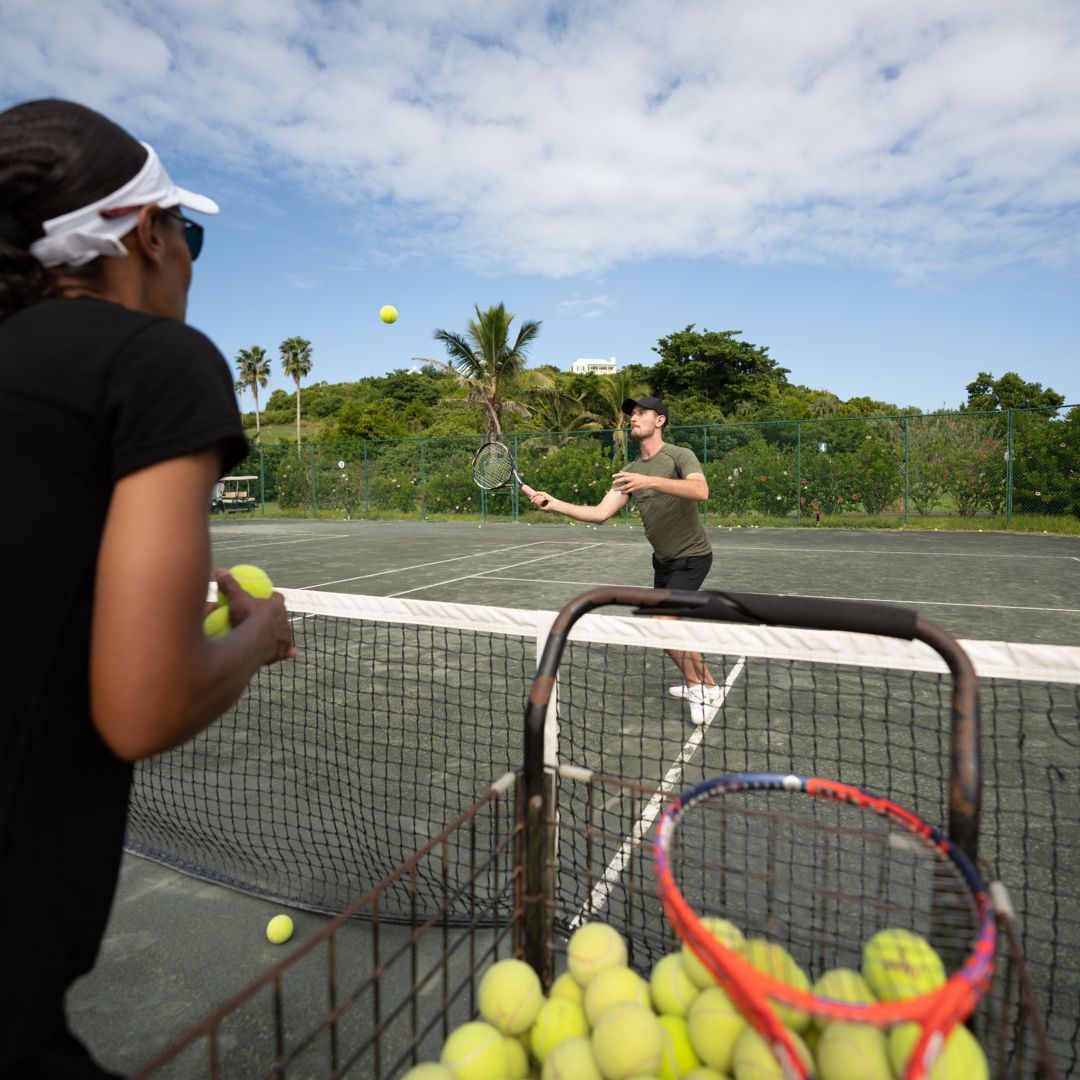 Tennis pro in foreground tossing tennis balls to practicing guest